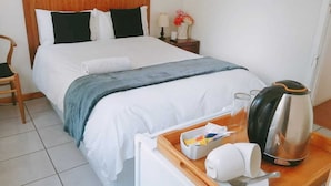 A neatly made bed with a wooden headboard, a bedside table with a lamp, a tray with a kettle, cups, and a spoon. - Steps Guest House (Gqeberha (Port Elizabeth))