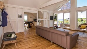 Amplia sala de estar con un sofá grande, piso de madera, una mesa de comedor con sillas, una zona de cocina y ventanas amplias que ofrecen vistas al exterior.
