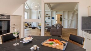 Cocina moderna con comedor, que incluye una mesa de madera con comida y una copa de vino, y vistas al salón con un sofá y una lámpara de araña.