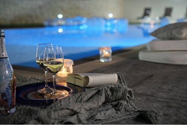 Una copa de vino blanco sobre una bandeja, una vela y un libro en una mesa cerca de una piscina.