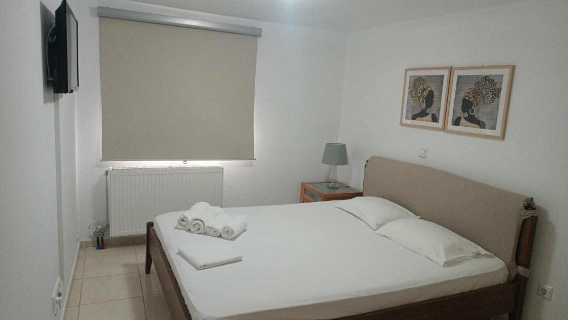 A bedroom with a bed, a nightstand with a lamp, a radiator, a window with blinds, and two wall-mounted pictures.
