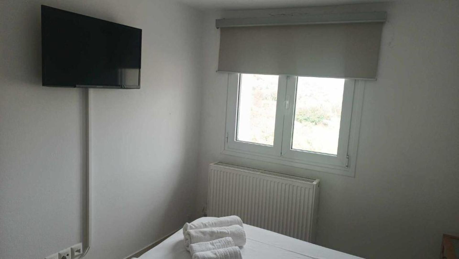 A hotel room with a bed, a TV mounted on the wall, a window with a blind, and a radiator.