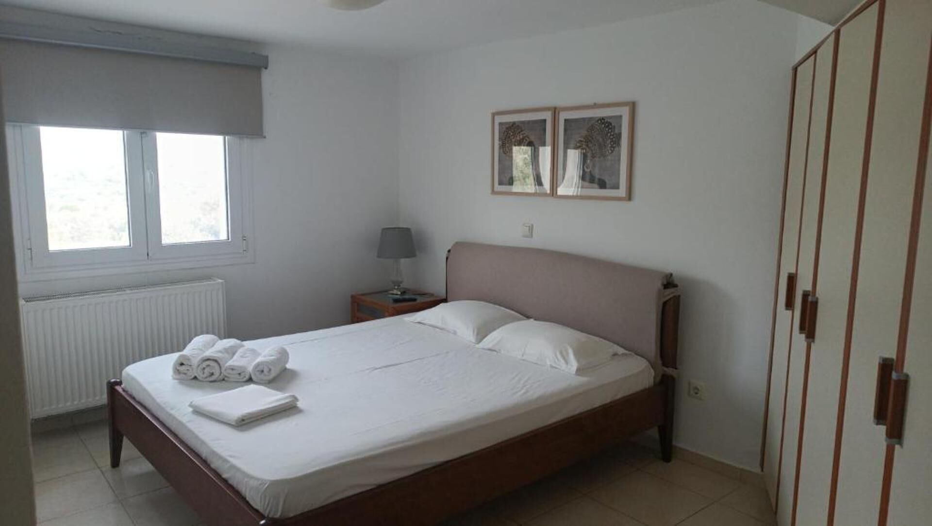 A neatly made bed with white linens and rolled towels, a bedside table with a lamp, and a window with blinds.