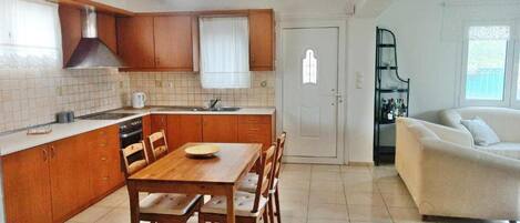A compact kitchen with wooden cabinets, a dining table with chairs, a built-in oven, and a sink. There is a white door and a black shelf with bottles.