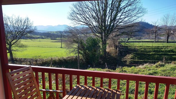 A balcony with a wooden table and two chairs, overlooking a green landscape with trees and distant mountains.