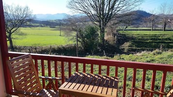 Een balkon met een houten tafel en twee stoelen, met uitzicht op een groen landschap met bomen en verre bergen.