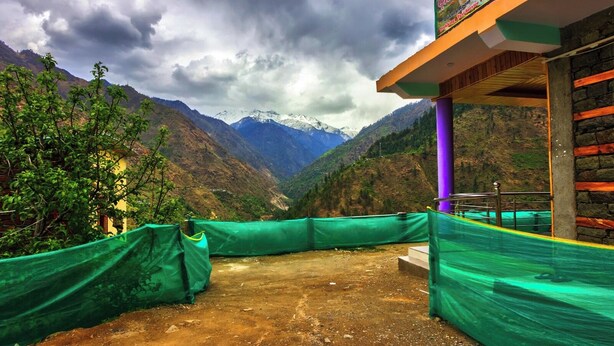 A mountain view with a building featuring a signboard, a purple pillar, and a green tarp-covered area. - Tirthan Height - Facing toward Snow View (Banjar)