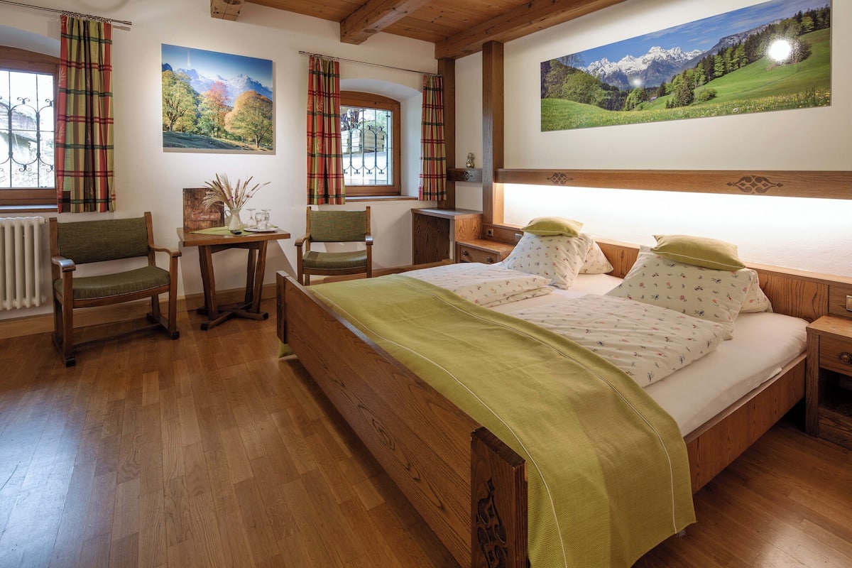 Una cama de madera con una colcha verde, dos sillas, una mesita y una ventana con cortinas.