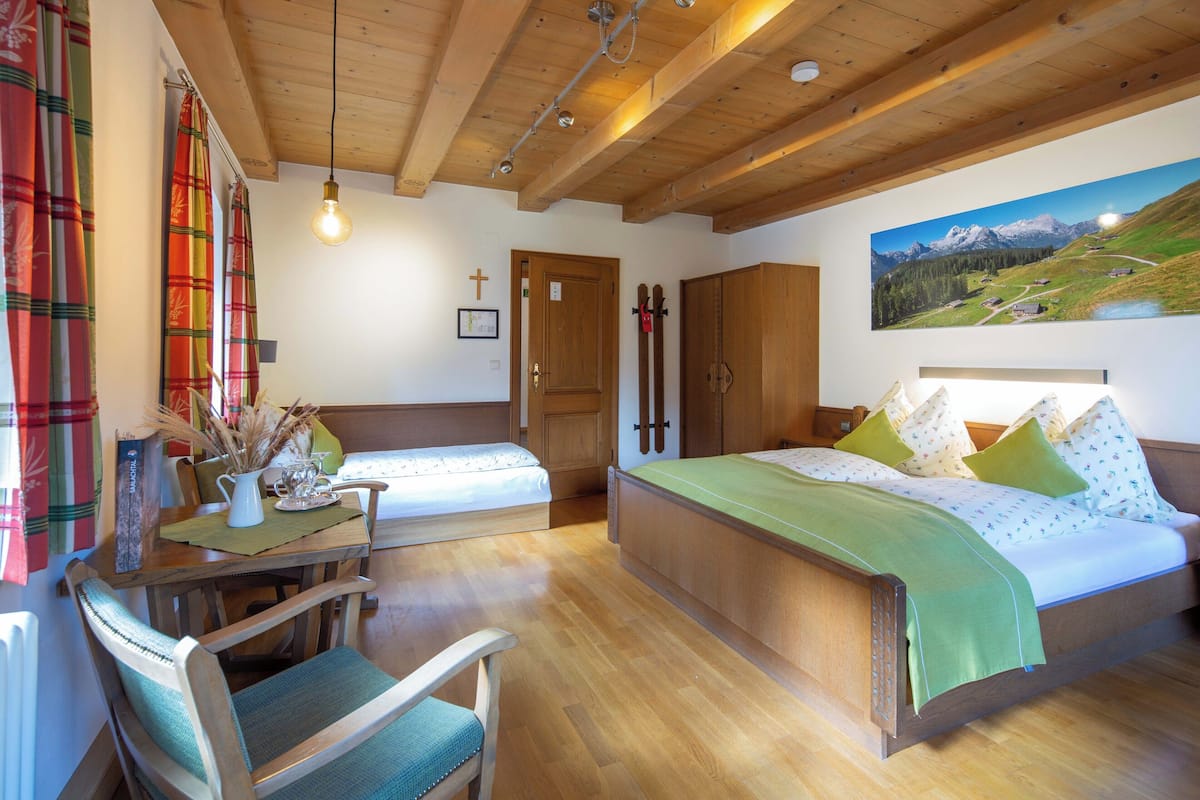 Un dormitorio con techo de madera, una cama con ropa de cama verde, una mesa de madera con un jarrón y un cuadro de un paisaje montañoso.