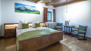 Ein Schlafzimmer mit einem hölzernen Bett, einem kleinen Tisch mit Stühlen, einem Fenster mit Vorhängen und einem Gemälde an der Wand.