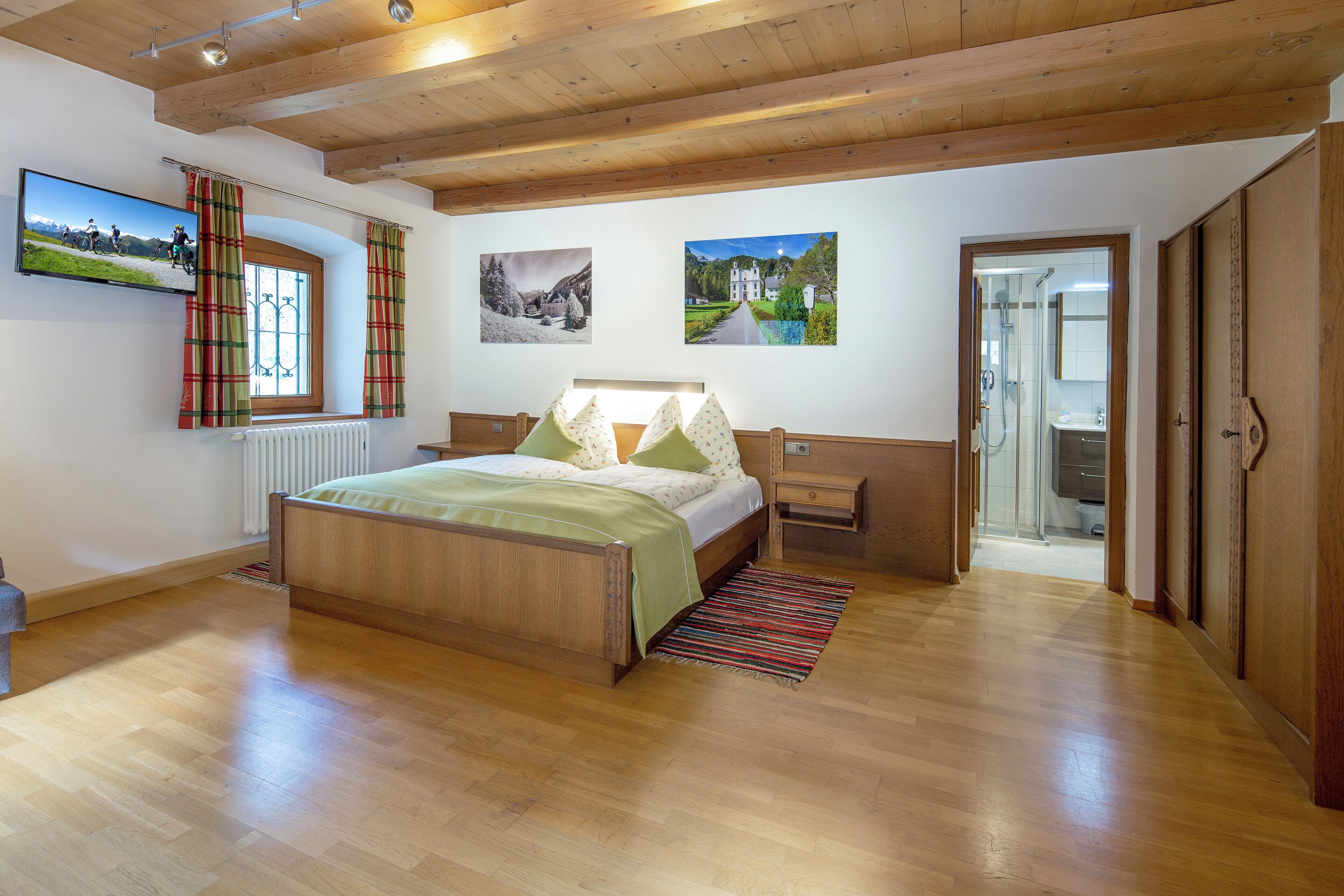 Un dormitorio con cama de madera, una mesita de noche, una ventana con cortinas y un televisor fijado a la pared.