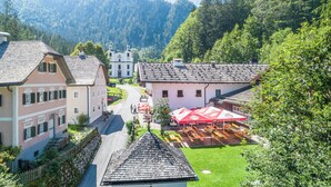 Ein traditionelles europäisches Dorf mit einem zentralen Gebäude, das eine rote und weiße Markise hat, umgeben von üppigem Grün und einem Bergpanorama.