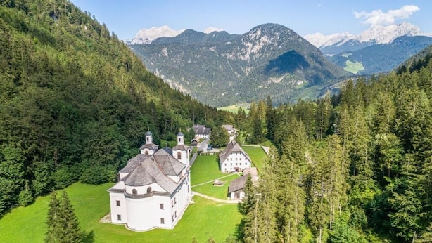 Ein großes, weißes, mehrstöckiges Gebäude mit einem runden Turm, umgeben von üppigem Grünwald und Bergen im Hintergrund.