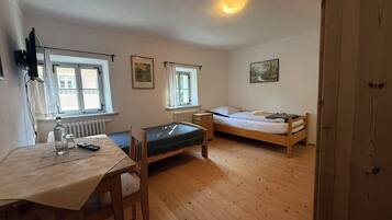 A small, well-lit room with a wooden floor, a single bed, a table with a chair, and two windows.