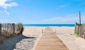 Une promenade en bois mène à une plage de sable fin, sous un ciel bleu et dégagé.