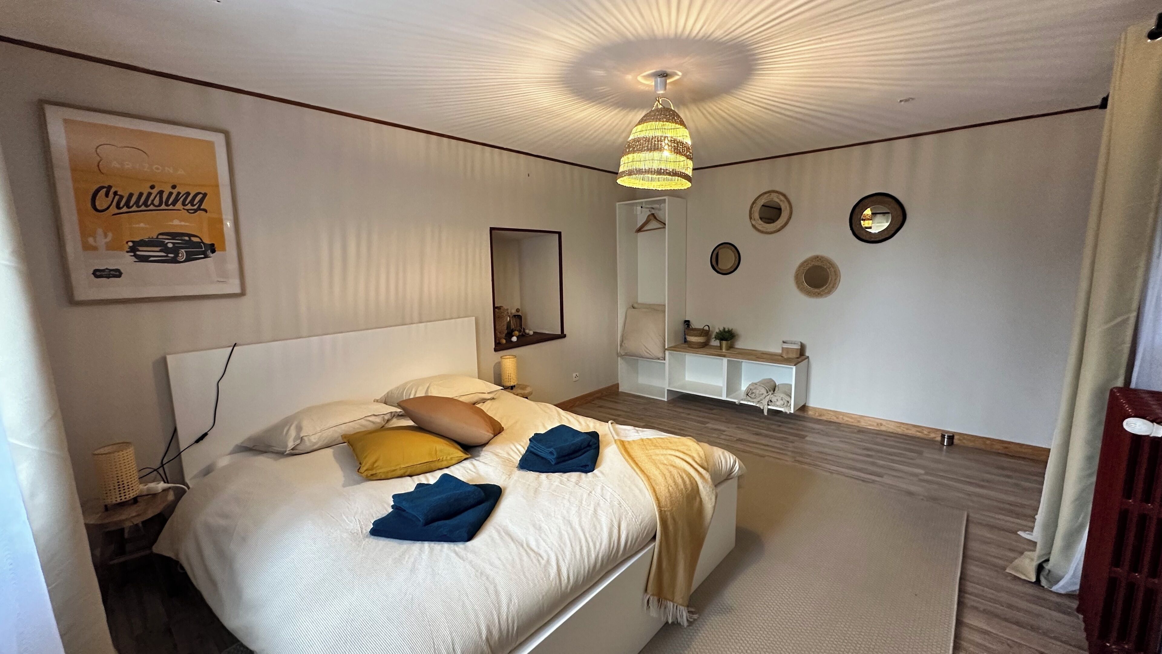 Ein ordentlich eingerichtetes Schlafzimmer mit einem großen Bett, einem Nachttisch, einer Hängelampe, einem Spiegel und einem an der Wand befestigten Regal mit Dekorationselementen.