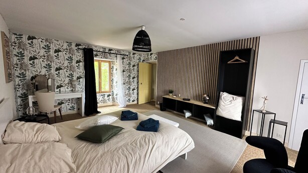 A bedroom with a large bed, a desk, a chair, a hanging light fixture, a wardrobe, and a mirror.