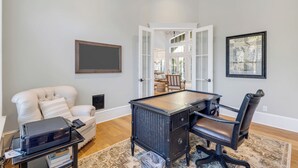 A home office with a desk, chair, printer, and a view into a living area through an open door.
