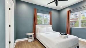 Een slaapkamer met een bed, een plafondventilator, een raam met gordijnen en een klein nachtkastje.