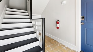 A staircase with black steps and white walls, a blue door, and a fire extinguisher mounted on the wall. - Red Stick Proud - 1BR Apartment, 2nd Floor (Baton Rouge)