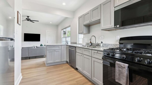 A modern kitchen with grey cabinets, a black stove, and a grey sofa in the adjacent living area. - Red Stick Proud - 1BR Apartment, 2nd Floor (Baton Rouge)
