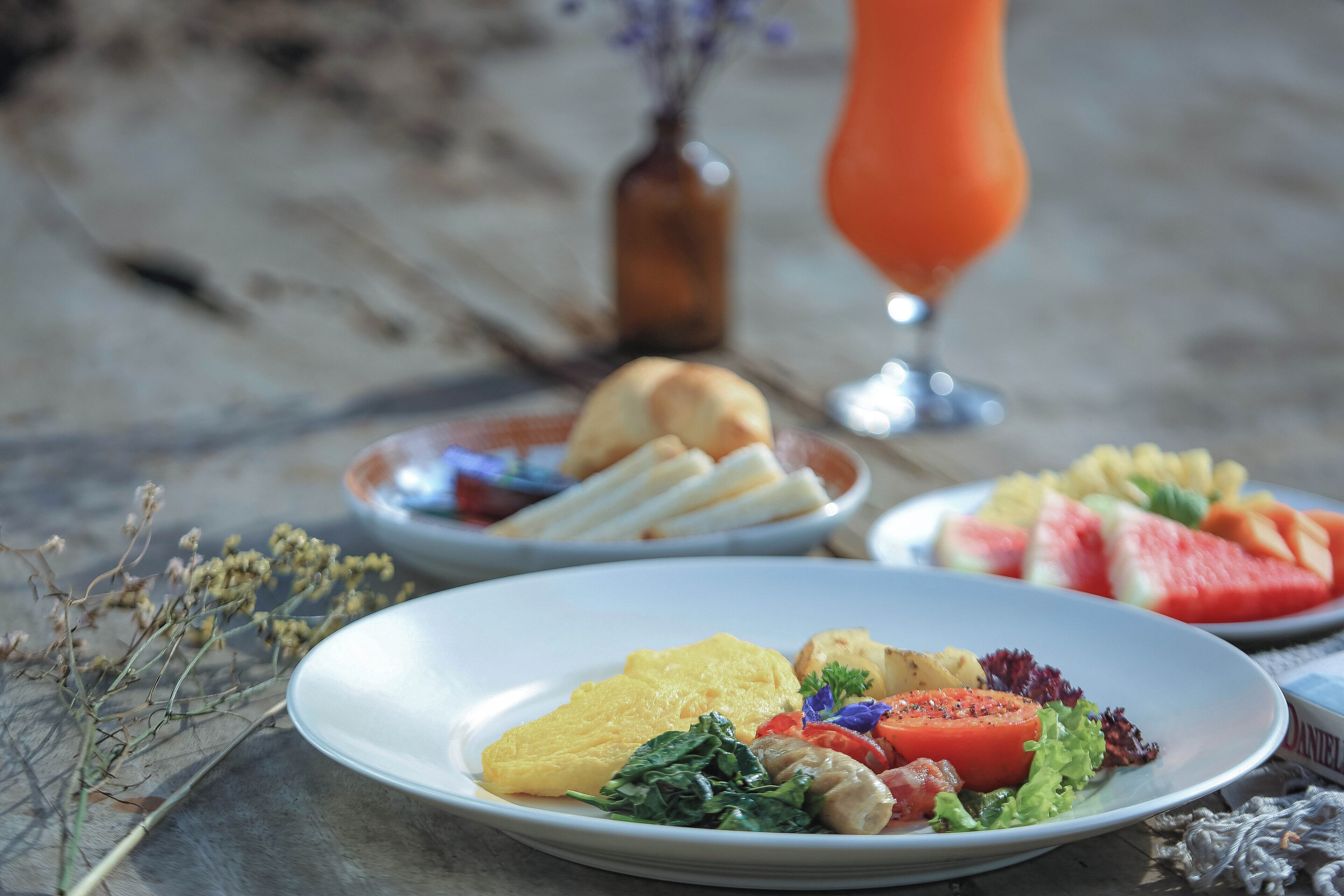 A table with plates of food, a glass of orange juice, and a vase with dried flowers.