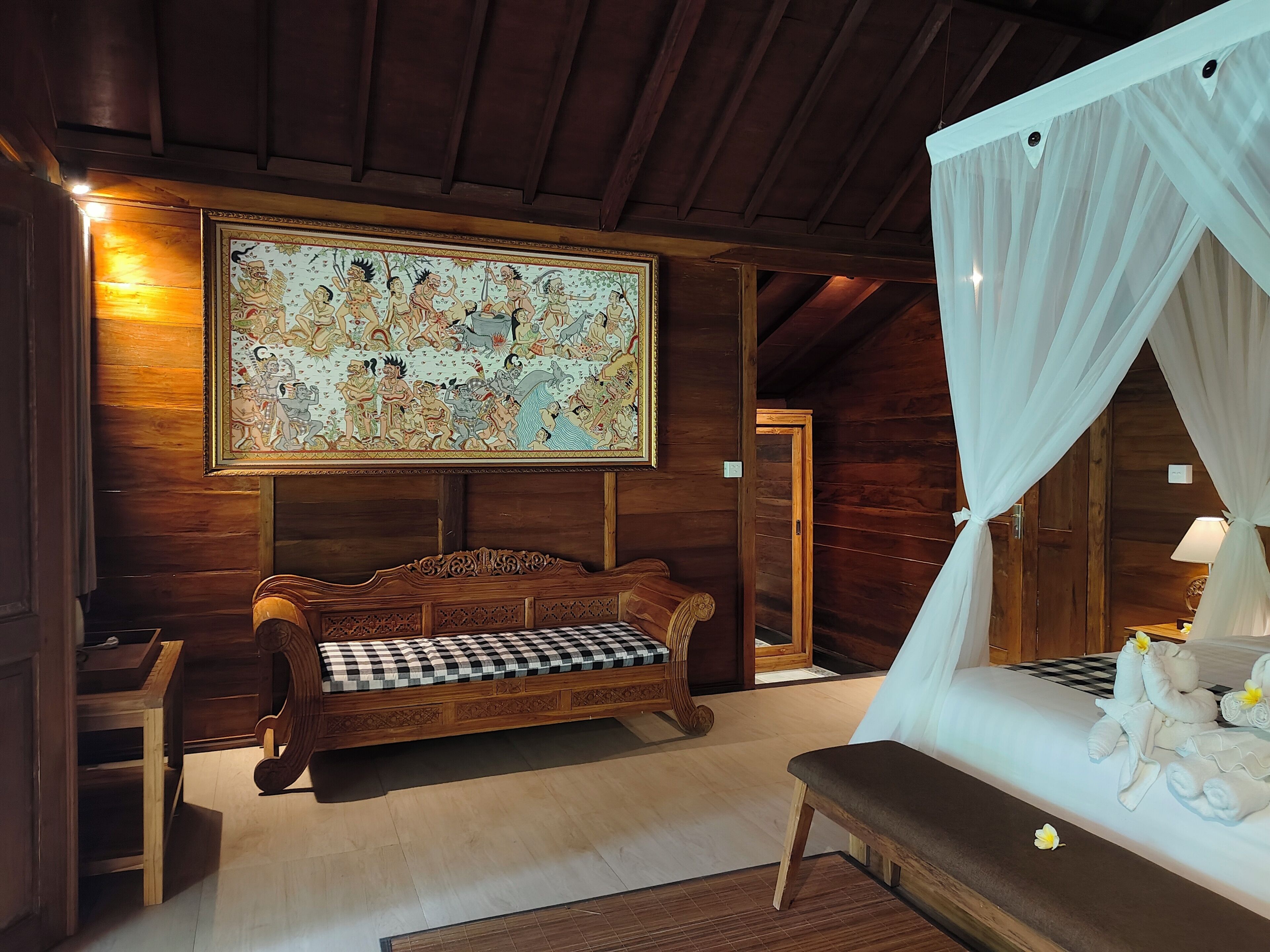 Interior de una cabaña de madera con un mural grande, un sofá a cuadros, una cama con sábanas blancas y una cama con dosel.