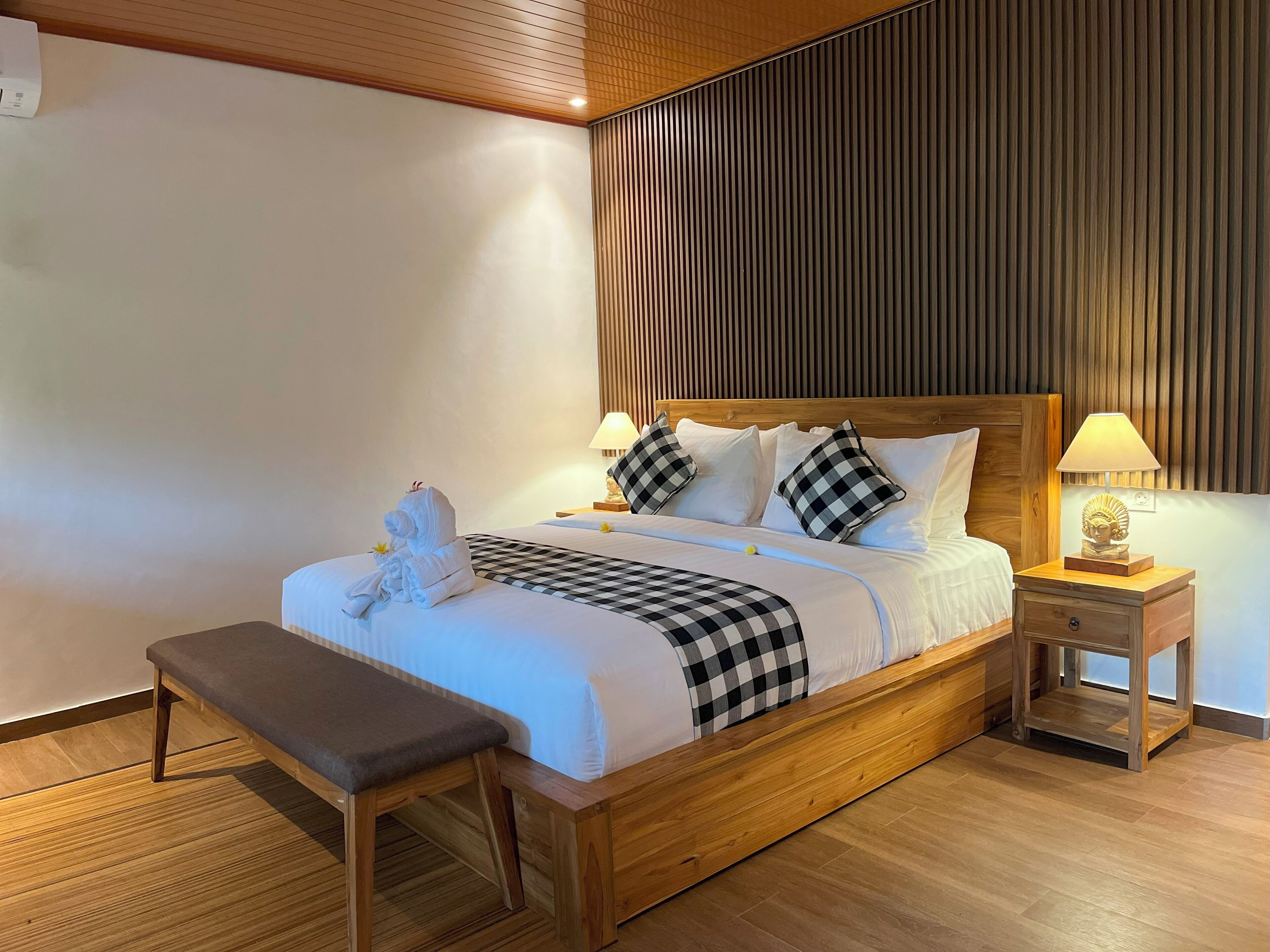 Een opgemaakt bed met geruit kussens, een bedlampje en een bankje.
