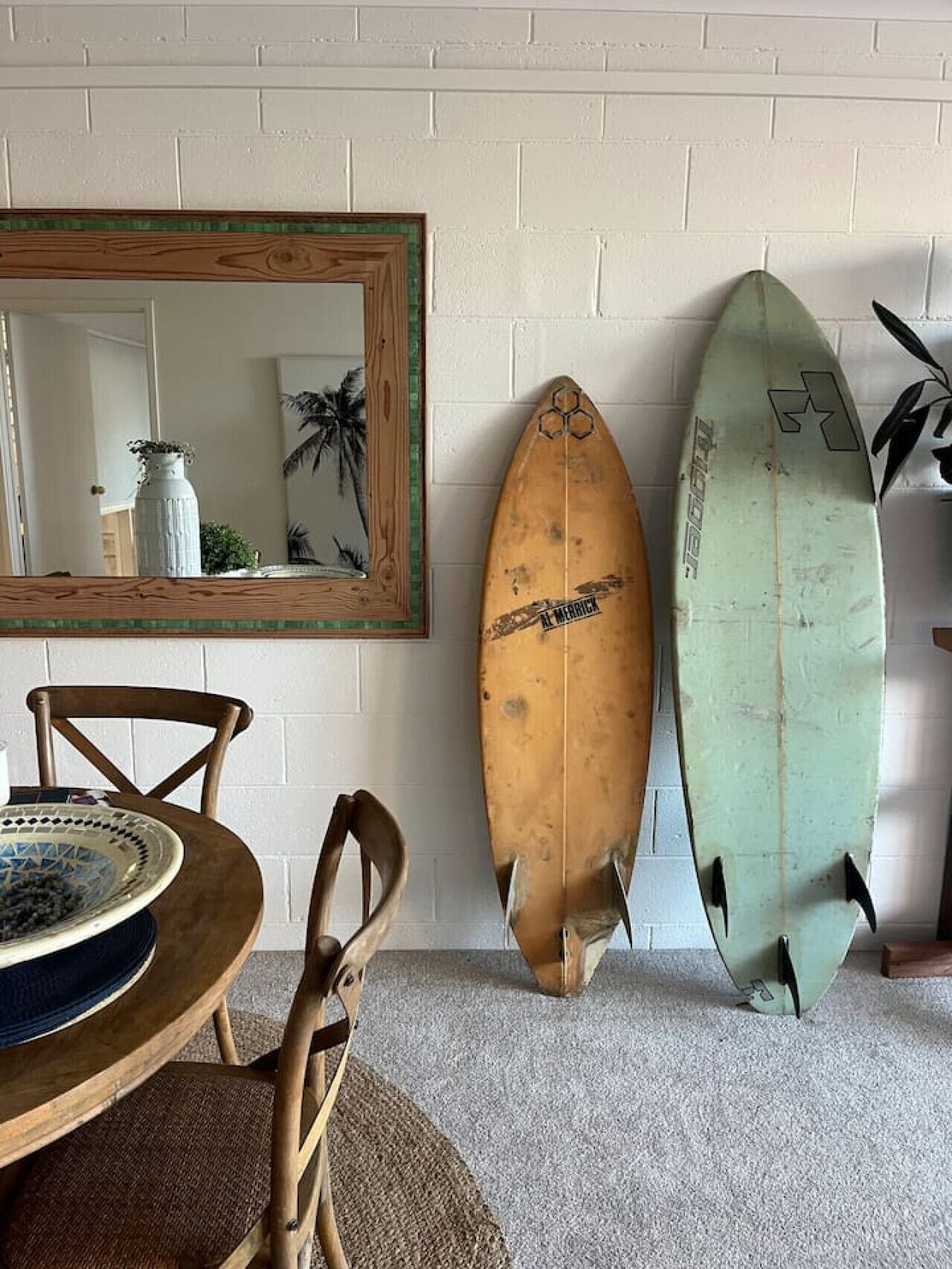 Una habitación con una mesa de comedor, sillas, un espejo y dos tablas de surf apoyadas contra la pared.