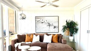 A living room with a brown sectional sofa, a small white table, and a ceiling fan. There is a framed picture on the wall and a potted plant on a side table.