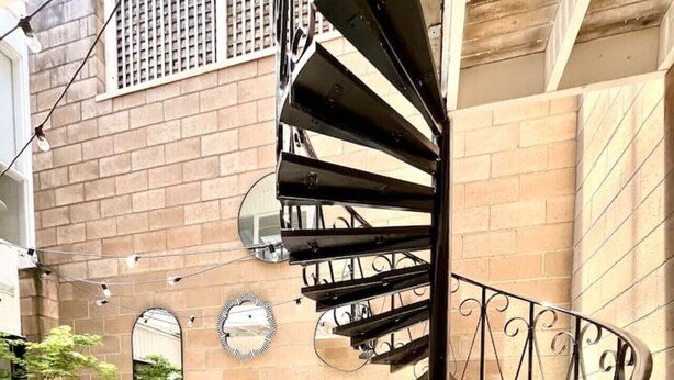 A spiral staircase with a metal railing, a bench, and potted plants in a courtyard with brick walls and a tiled floor.