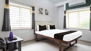 A bedroom with a bed, two windows with curtains, a bedside table with bottles, and an air conditioning unit. - Hotel Crystal INN (Bhubaneshwar)