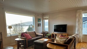 Ein gemütliches Wohnzimmer mit einer Couch, Hocker und Couchtisch, einem Flachbildfernseher und Blick ins Freie.