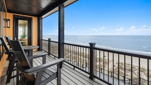 Um deck de madeira com cadeiras e uma mesa, com vista para uma praia e céu claro.