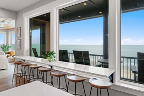 A modern kitchen with a large window overlooking the ocean, bar stools, and a dining table.