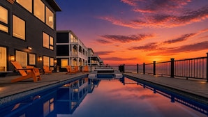 Piscina com vista para o pôr do sol, cadeiras de deck e um edifício com amplas janelas.