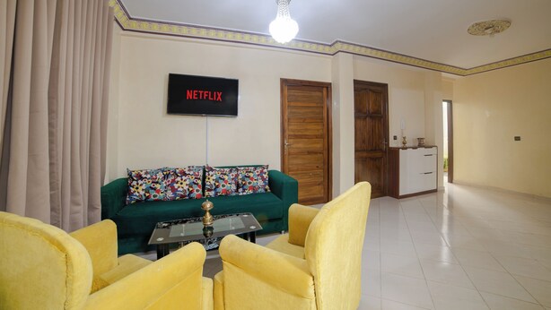 A living room with a green sofa, yellow armchairs, a coffee table, and a flat-screen TV displaying "NETFLIX".