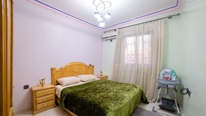A bedroom with a bed, wooden nightstand, green bedspread, and a baby high chair.