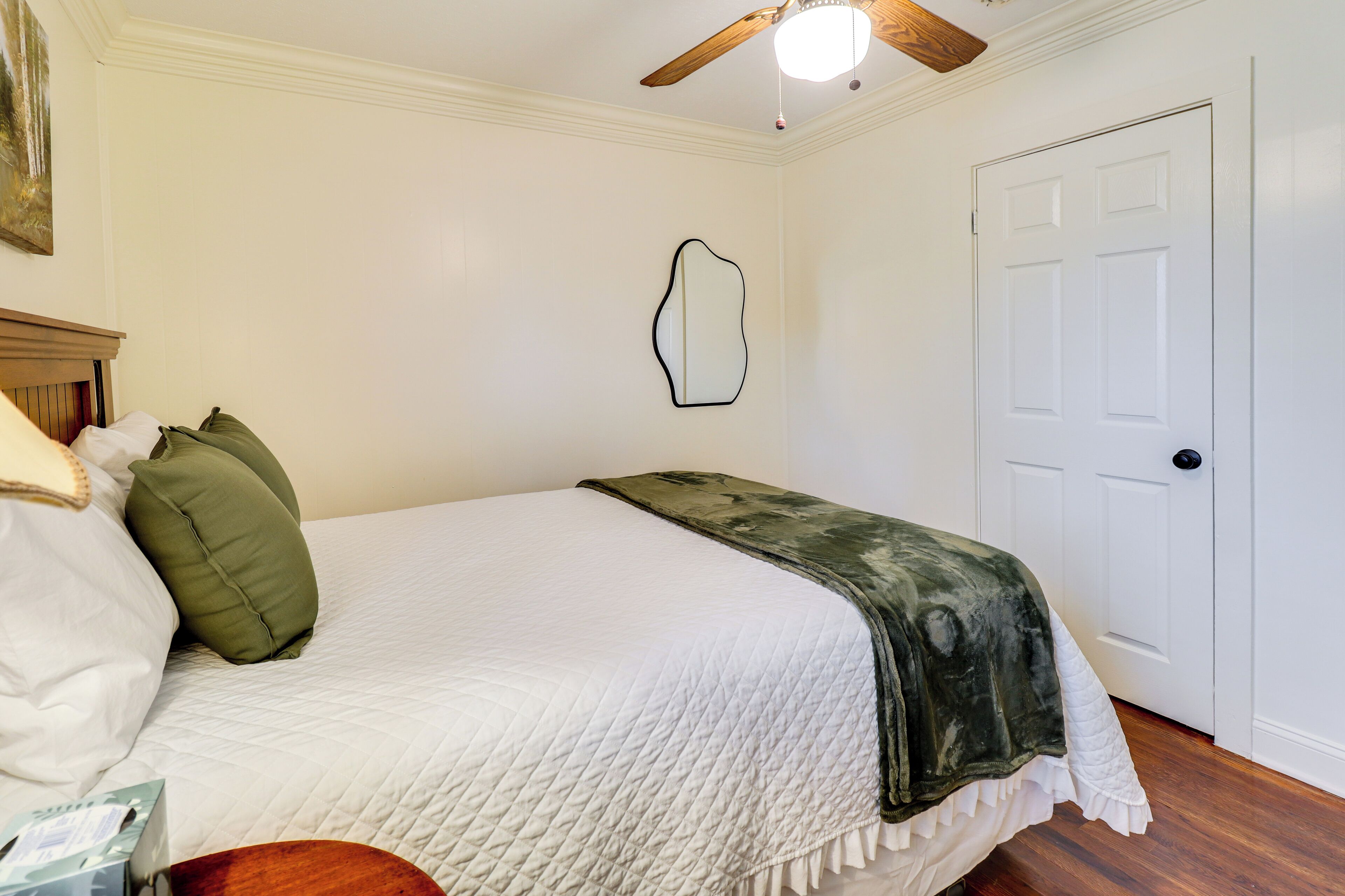 A bedroom with a bed, a ceiling fan, a door, and a wall-mounted mirror.