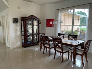 A dining room with a large wooden table, six chairs, a glass cabinet, and a view through a large window. - The ArtHouse - modern, stylishly furnished vacation home with pool! (Lehigh Acres)
