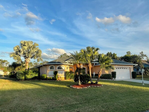 A single-story house with a garage, surrounded by a well-maintained lawn and palm trees. - The ArtHouse - modern, stylishly furnished vacation home with pool! (Lehigh Acres)