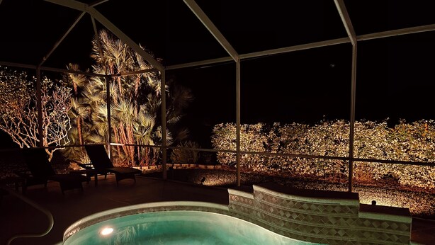 An indoor swimming pool with a tiled edge, surrounded by a covered patio area with chairs and a view of a landscaped garden at night.