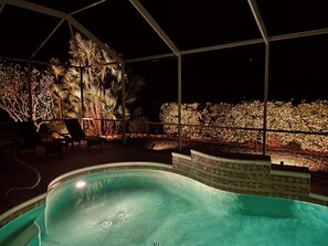 An indoor swimming pool with a tiled edge, surrounded by a covered patio area with chairs and a view of a landscaped garden at night. - The ArtHouse - modern, stylishly furnished vacation home with pool! (Lehigh Acres)