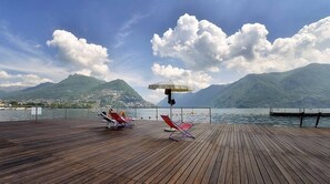 A wooden deck with two red lounge chairs and a white umbrella, overlooking a lake with mountains in the background.