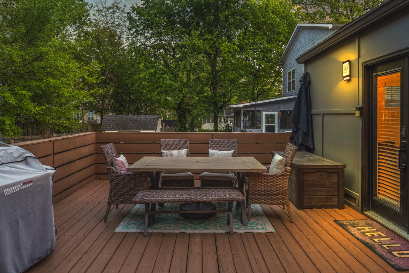 An outdoor deck with a wooden table and wicker chairs, surrounded by a wooden fence and trees.