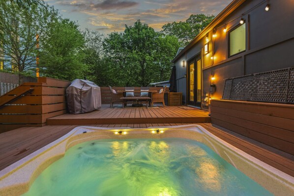 A hot tub on a wooden deck with outdoor furniture and a covered grill, surrounded by trees and a building with large windows.