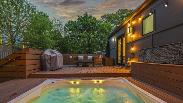 A hot tub on a wooden deck with outdoor furniture and a covered grill, surrounded by trees and a building with large windows. - Hot Tub | Fully-Fenced Yard |The Bubbly Suite (Atlanta)