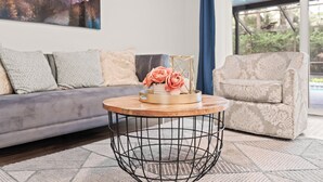 Een woonkamer met een grijze bank, een fauteuil met patroon, een ronde salontafel met een bloevas, en een groot raam met uitzicht op groen.