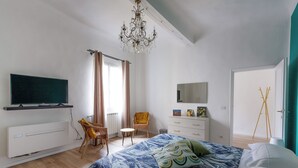 Une chambre à coucher comprenant un lit, une télévision, une commode, une chaise et une fenêtre avec des rideaux.
