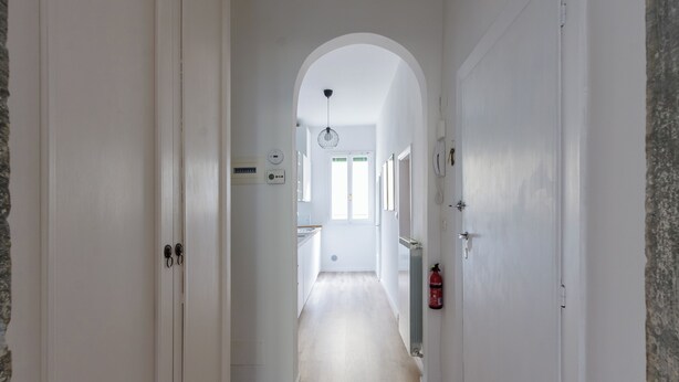 Un couloir étroit aux murs blancs et au plancher en bois, qui mène à un espace cuisine doté d’une fenêtre et d’une porte au fond.